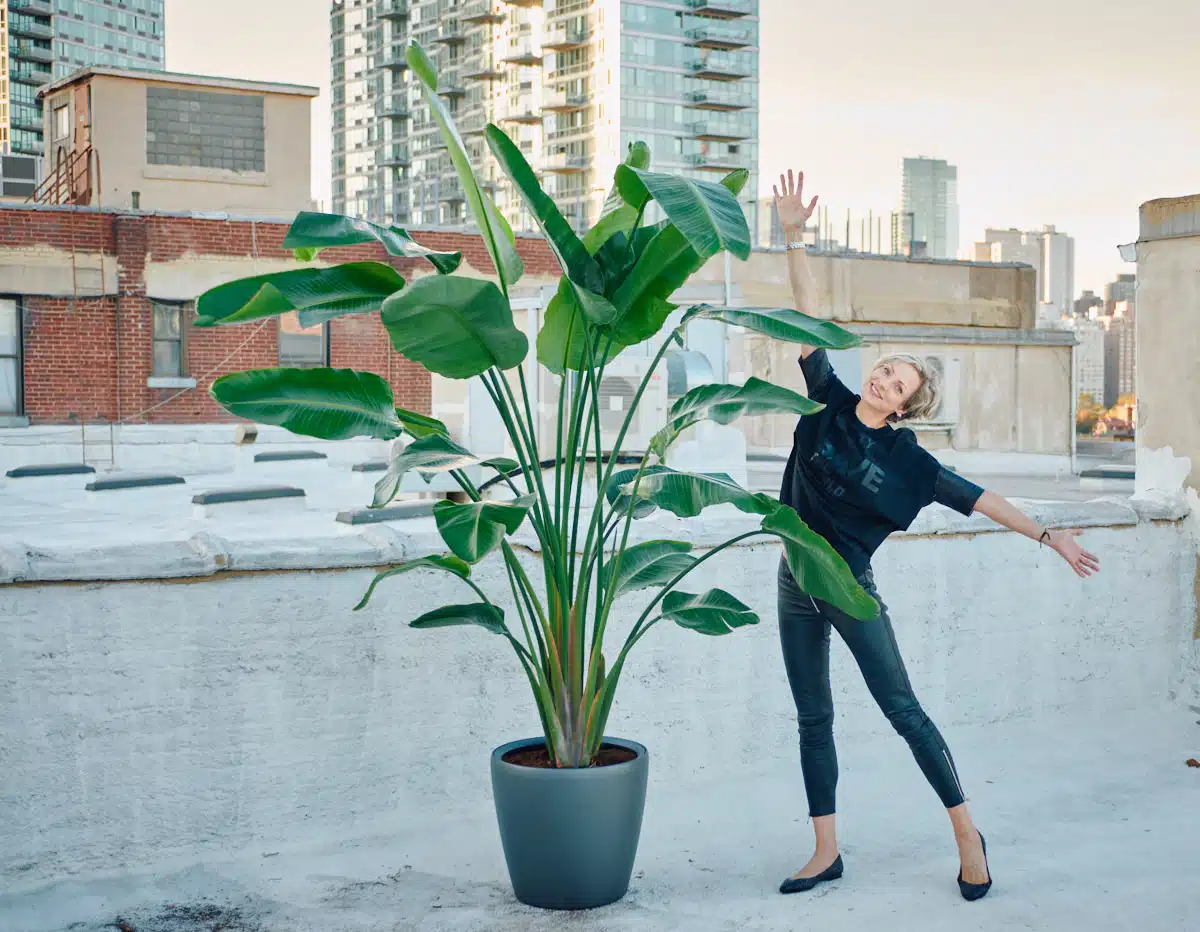 Juliette, the founder of My City Plants, playfully posing next to a large bird of paradise plant on an urban rooftop.