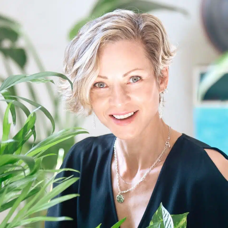 Smiling woman with short blonde hair next to indoor plants.