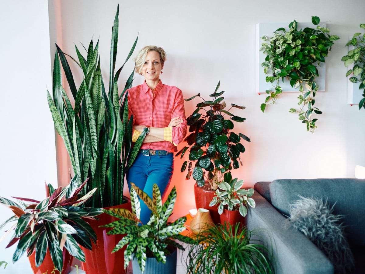 Confident businesswoman in stylish casual attire posing with a collection of indoor plants in a modern office lounge setting