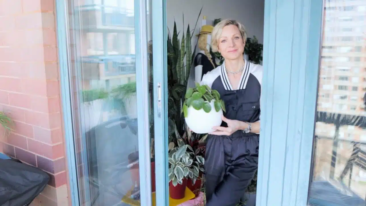 A smiling Juliette in a dark jumpsuit and gloves, standing in a glass-enclosed balcony, holding a spherical white planter with a green leafy plant. A cozy urban garden vibe emanates from the surrounding plants and the cityscape in the background.