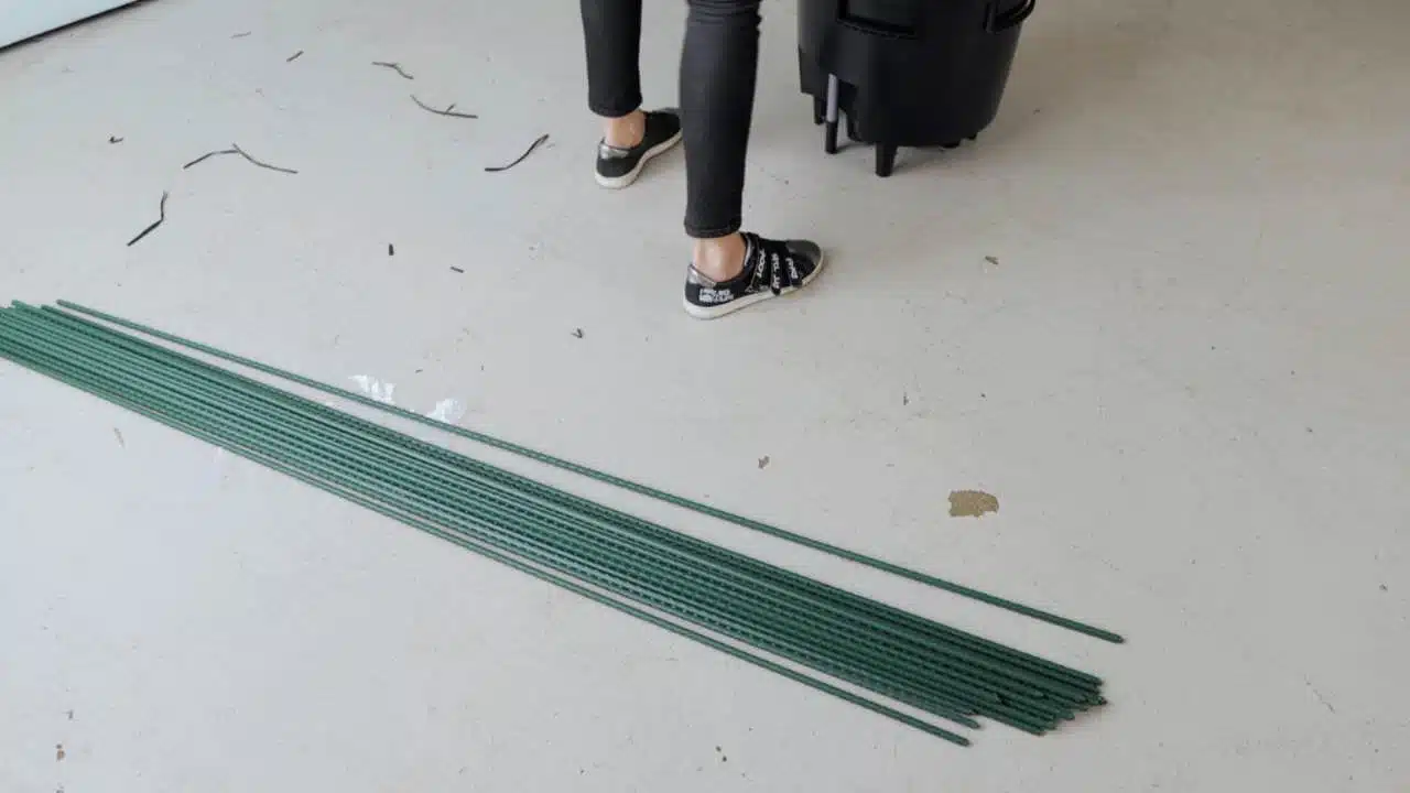 A collection of long green metal stakes lying on a concrete floor with a person's feet standing nearby, indicating preparation for garden work or plant support.